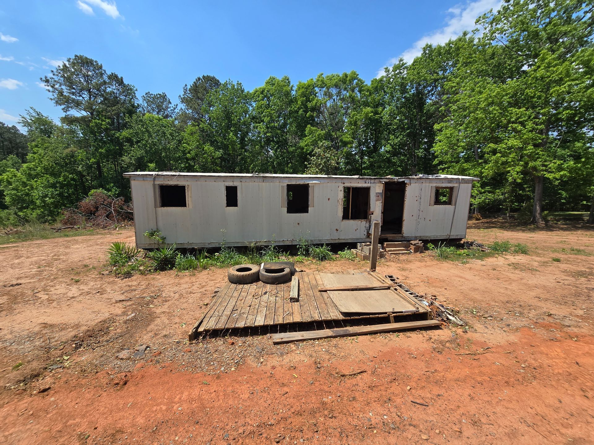 Abandoned white trailer in a clearing, with a dirt yard and trees under a blue sky