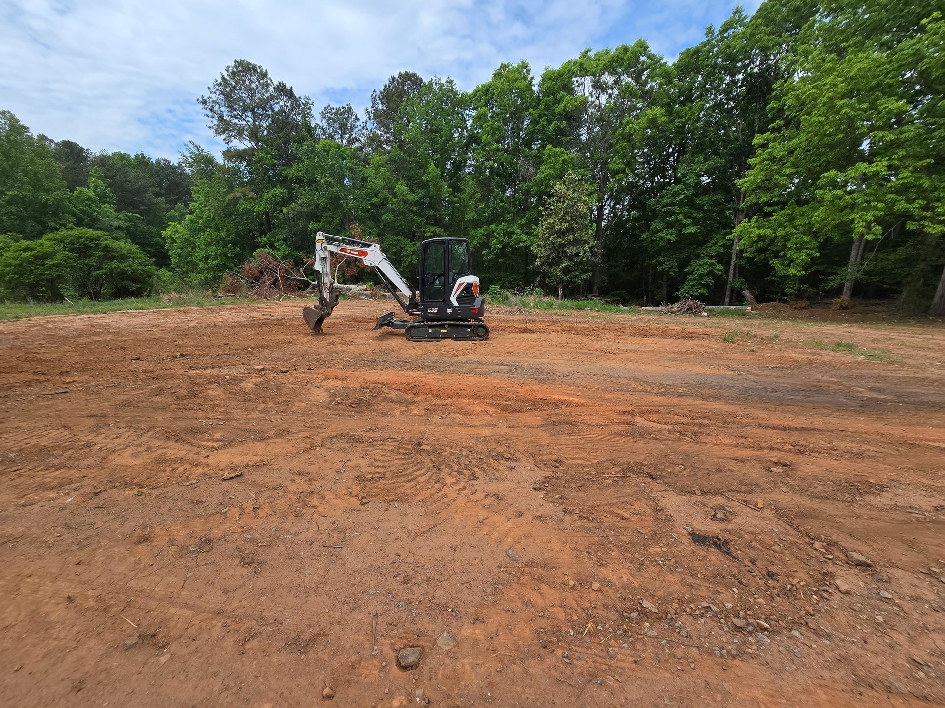 Excavator clearing a large dirt lot beside a tree line under a blue sky