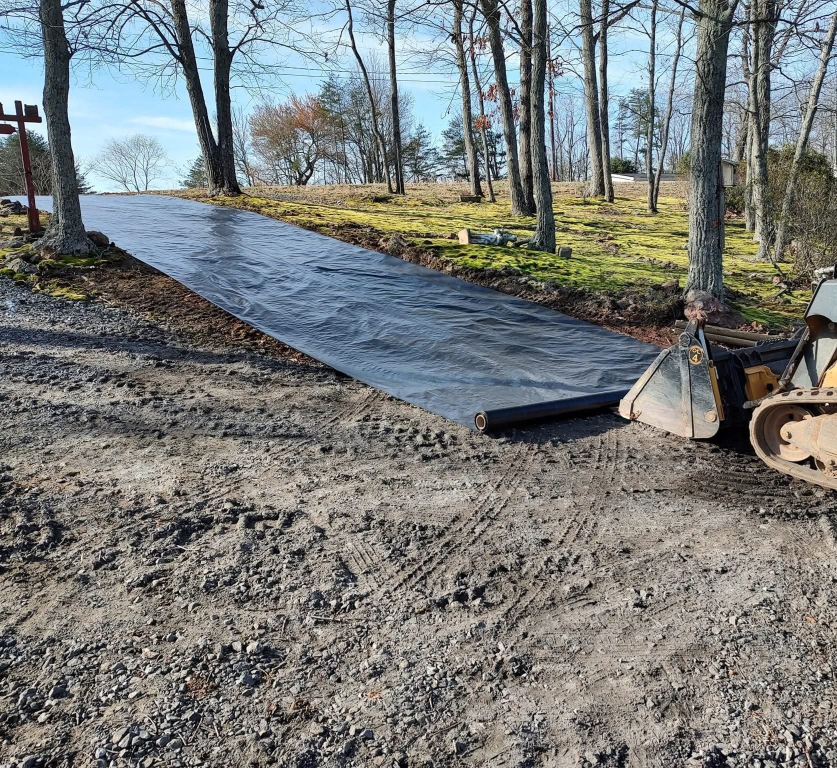 Freshly paved driveway under construction in a wooded area, with a skid steer and gravel foreground