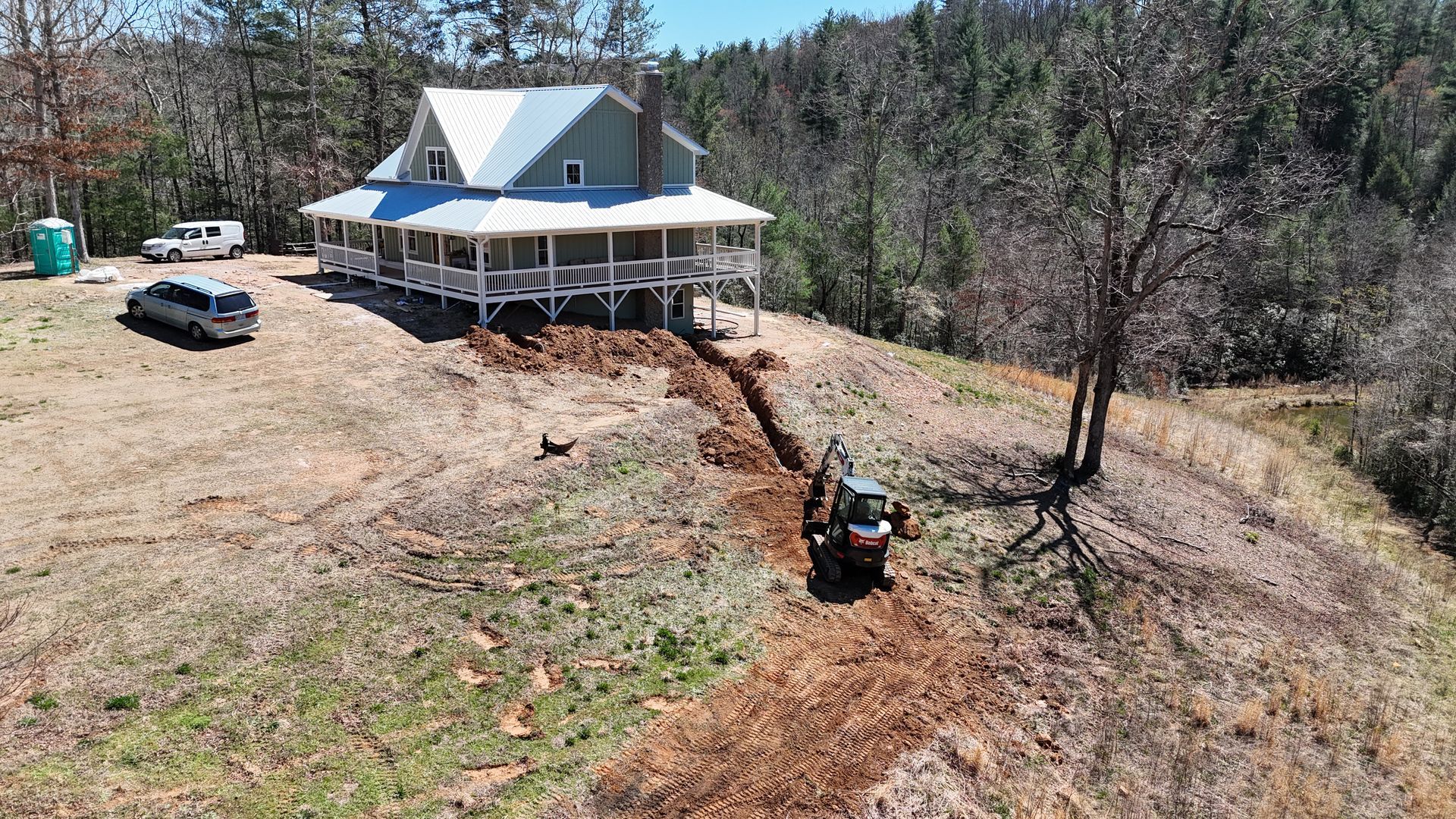 Rural house with a white porch on a hillside, surrounded by bare trees and a dirt driveway with a parked ATV.