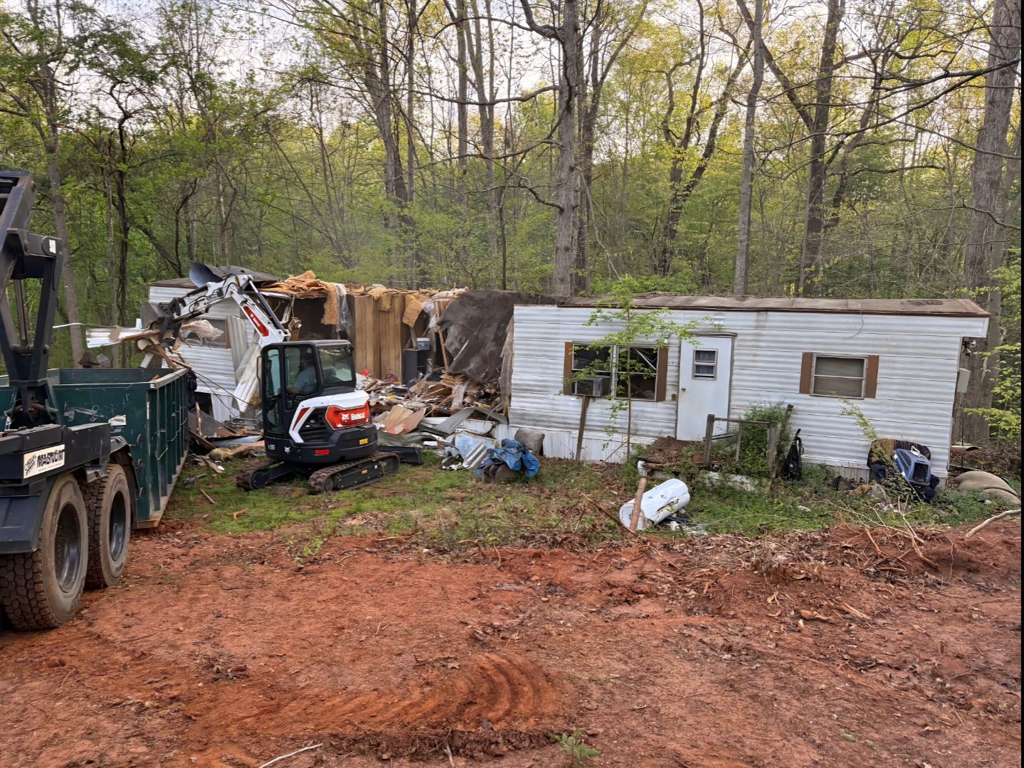 Bulldozer beside a cluttered white mobile home in a wooded yard with debris and dirt foreground