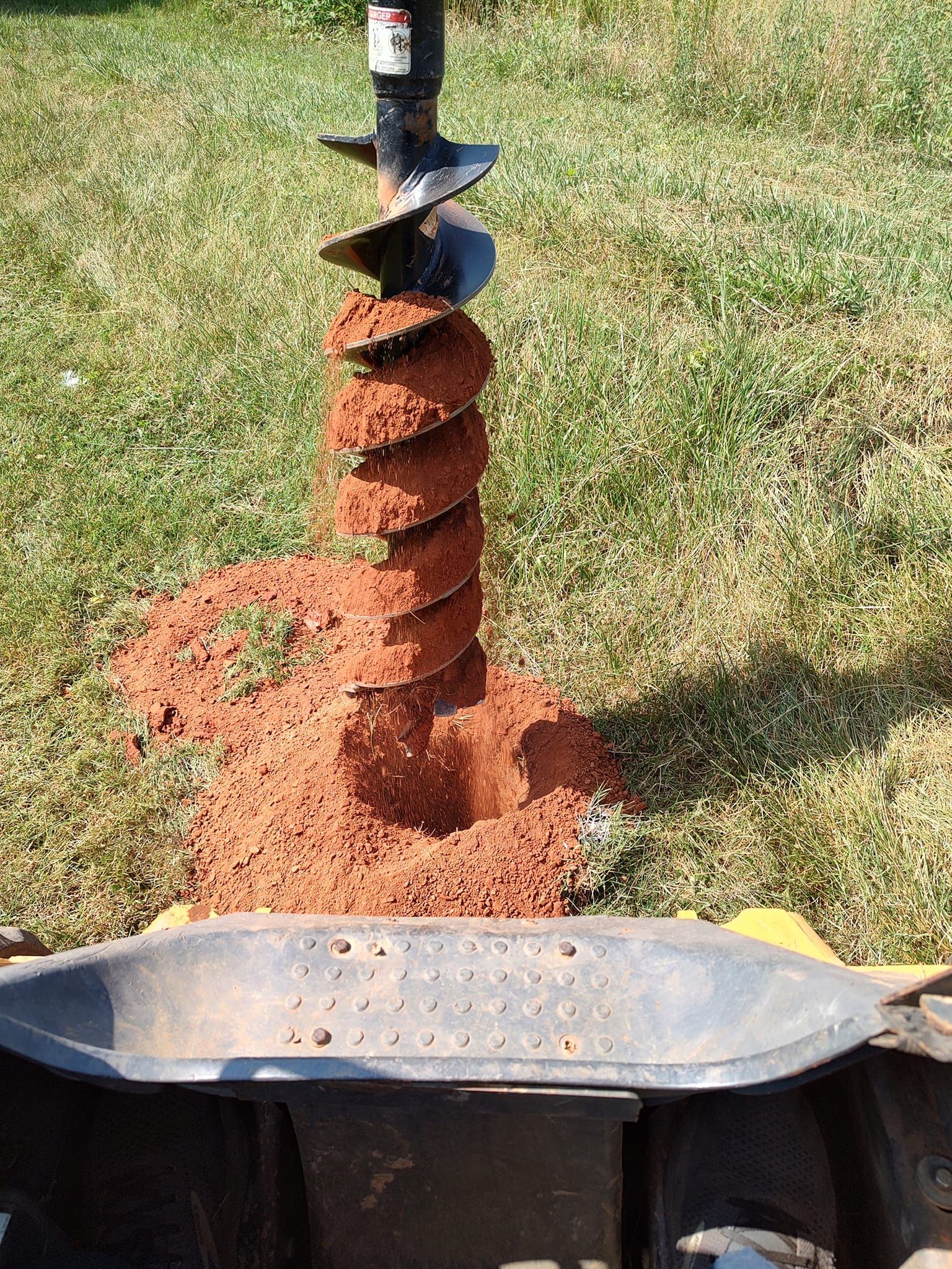 Rusty auger bit drilling red clay soil beside a black planter in a sunny yard