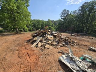 Demolition debris piled on dirt ground beside trees under a blue sky