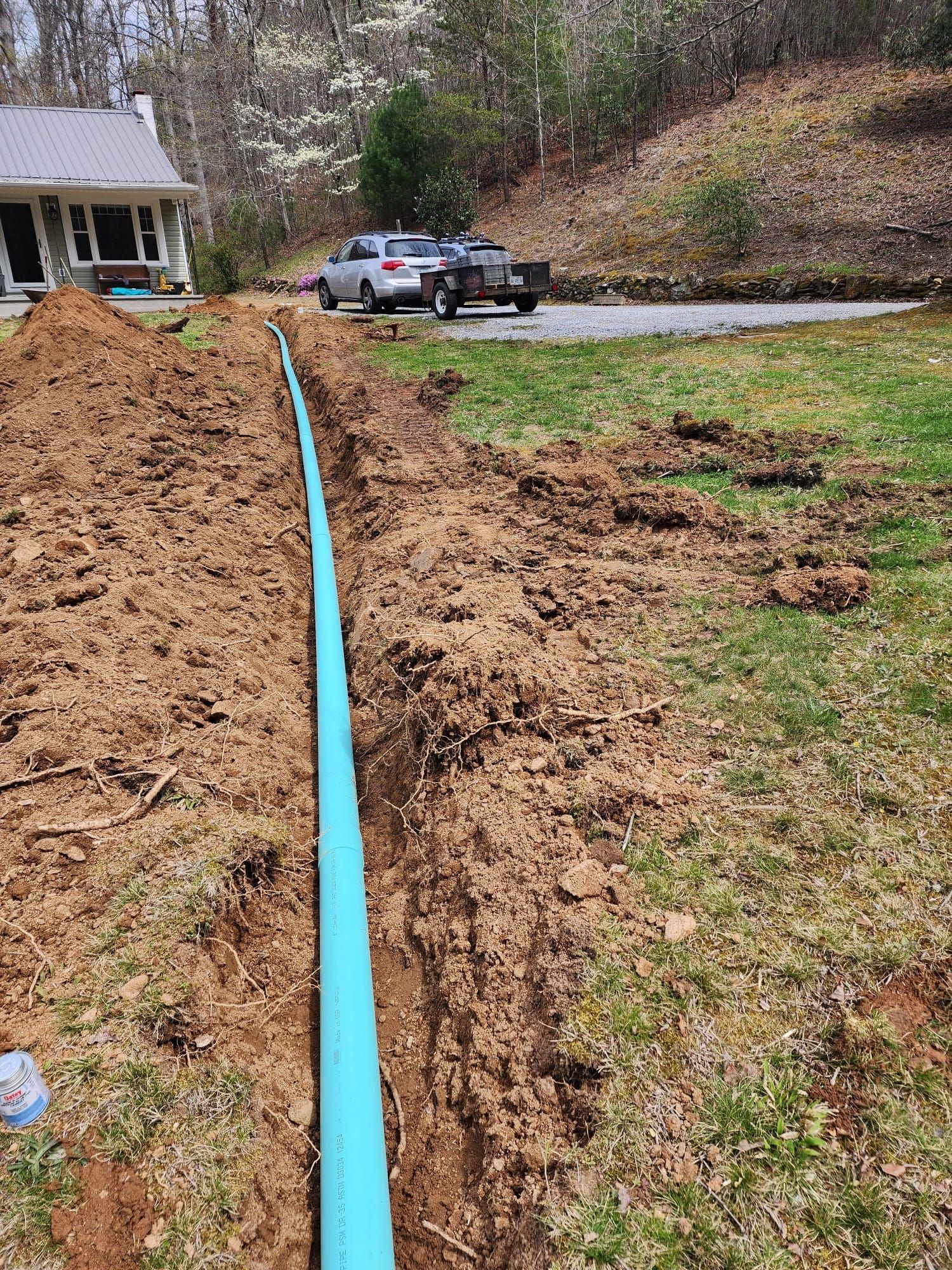 Blue utility pipe laid in a trench across a muddy yard near a house and parked cars.