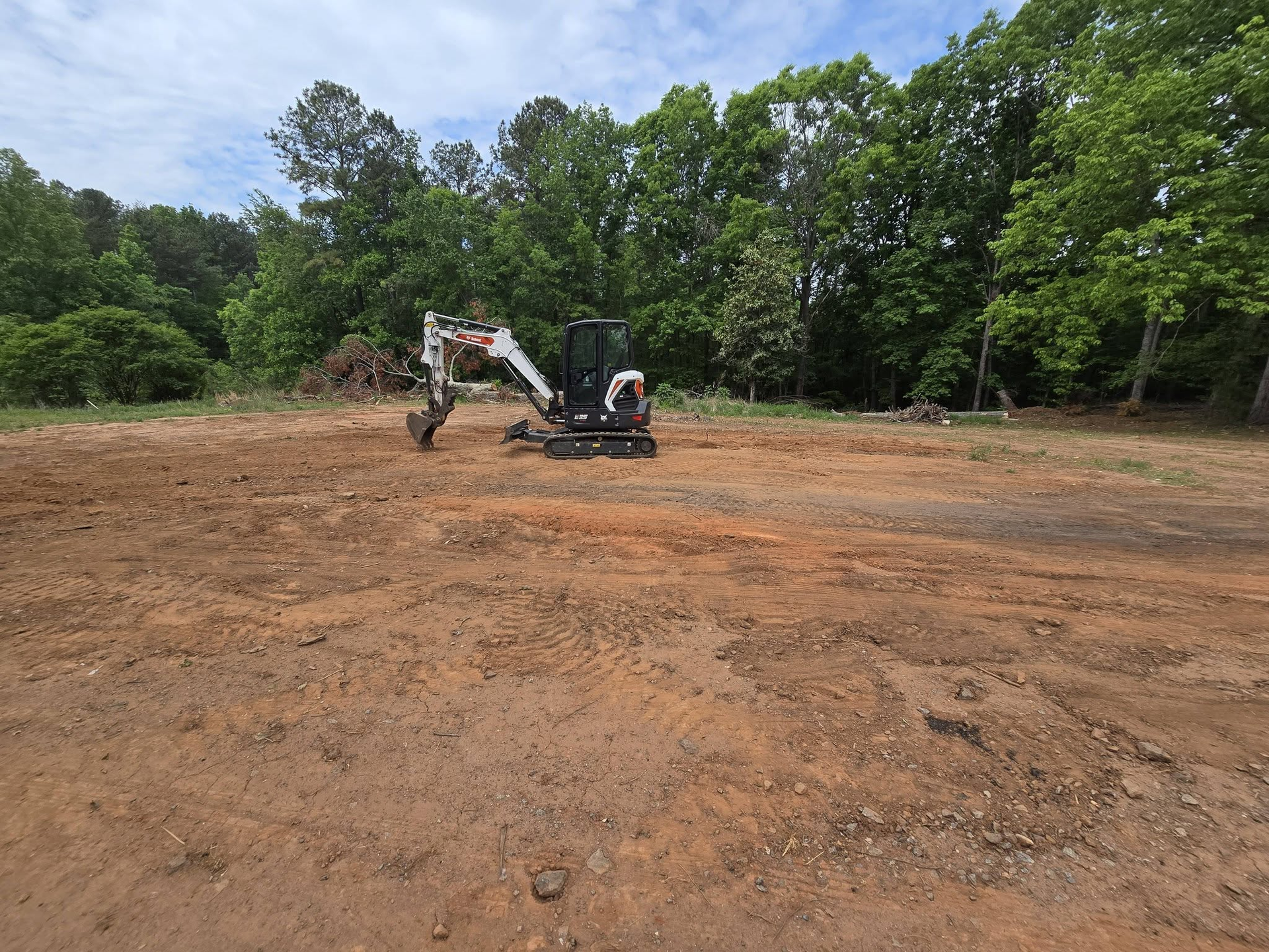 Excavator clearing a dirt lot with trees in the background under a bright blue sky