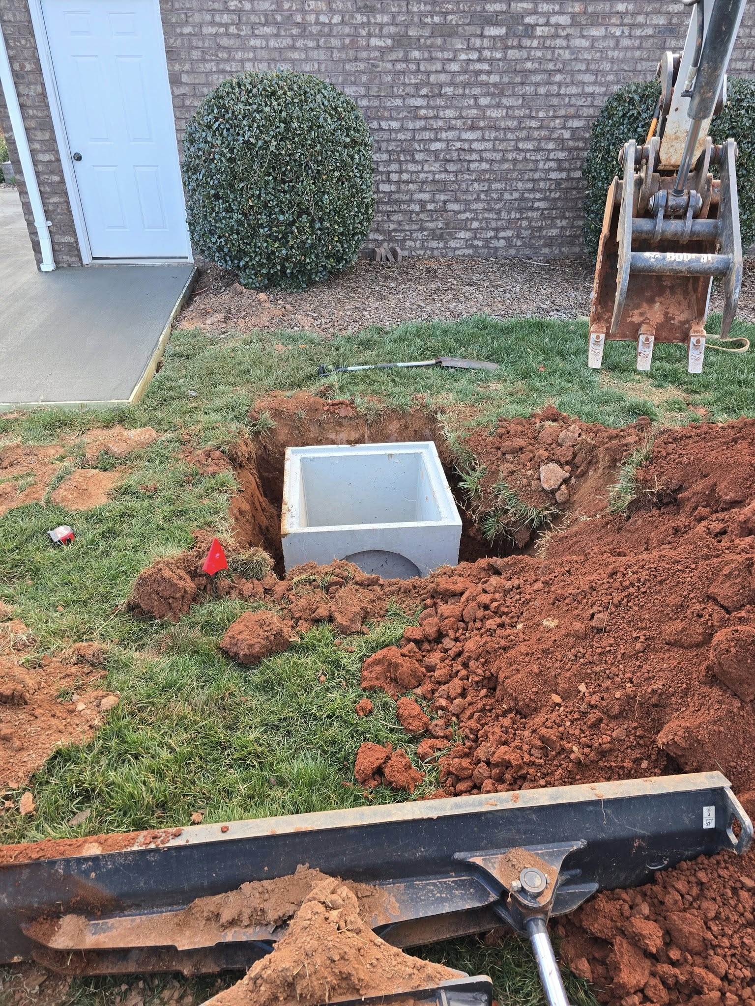 Backyard with a dug trench, exposed red soil, and a gray utility box near a brick wall.