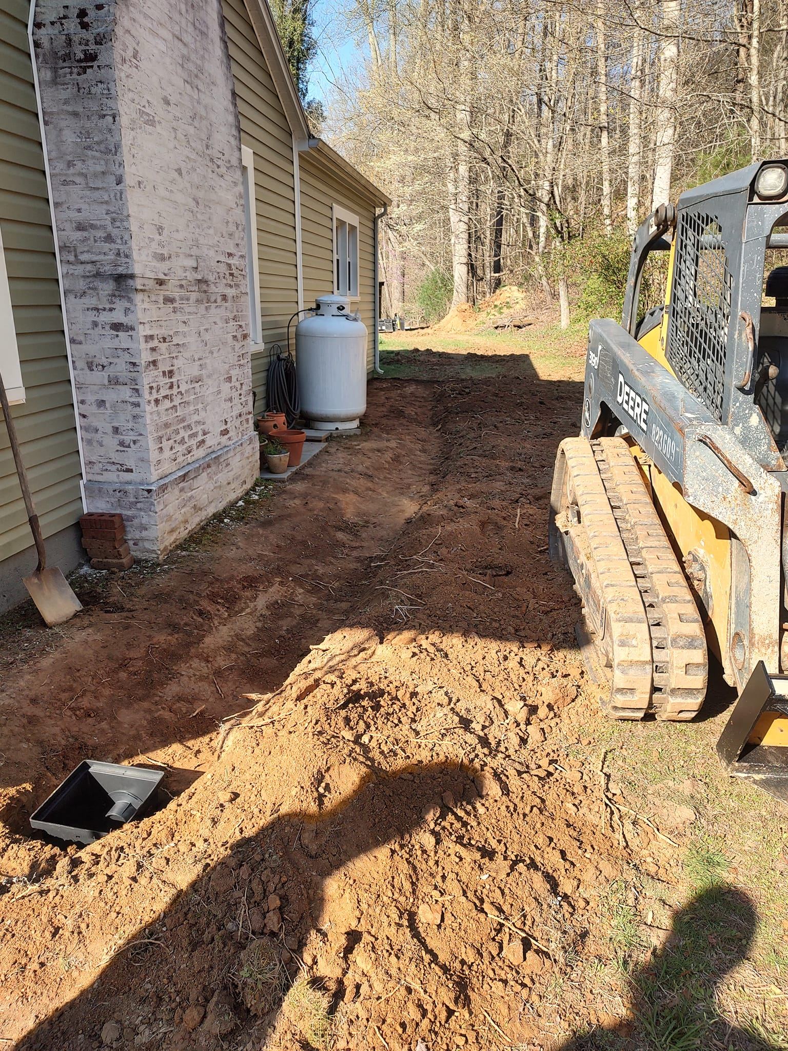 Backyard construction area with dirt, a mini excavator, house siding, and a propane tank.