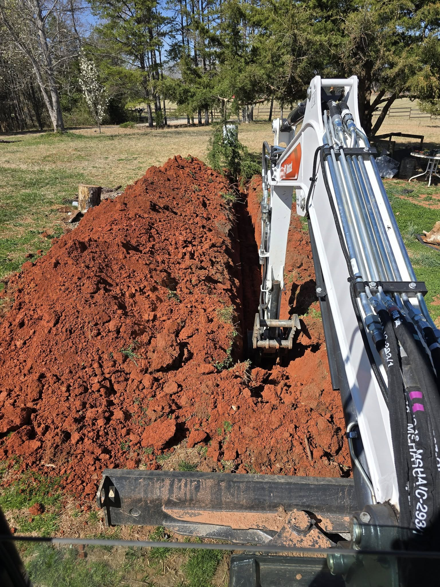 Excavator arm beside a large pile of red dirt in a grassy yard under trees