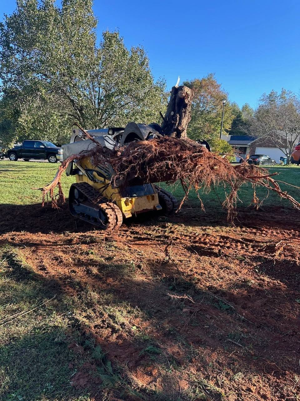 Yellow skid steer pushing a pile of dirt in a grassy yard with trees and parked cars nearby