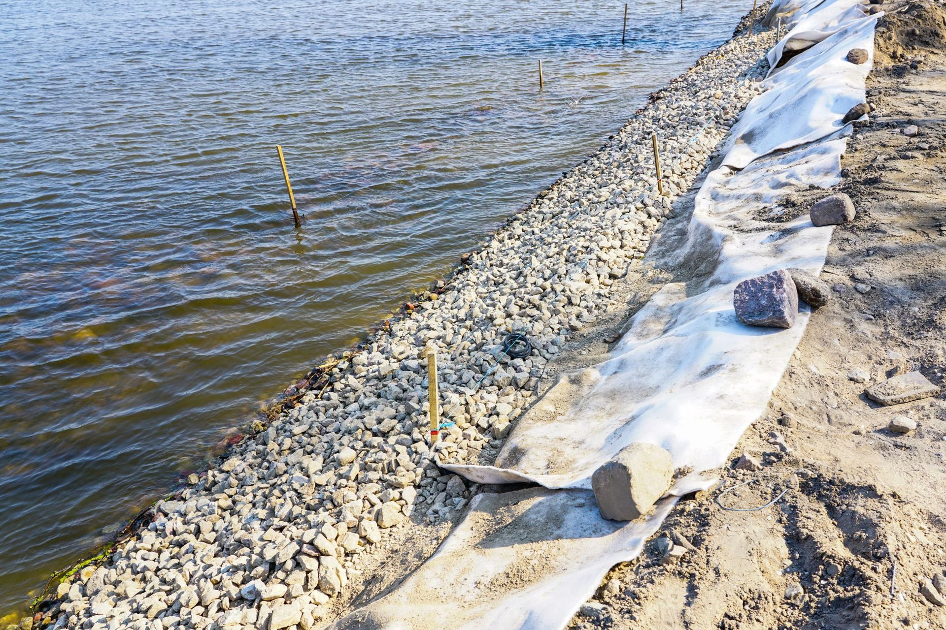Rock-lined shoreline with white erosion barrier beside rippling water and sandy bank