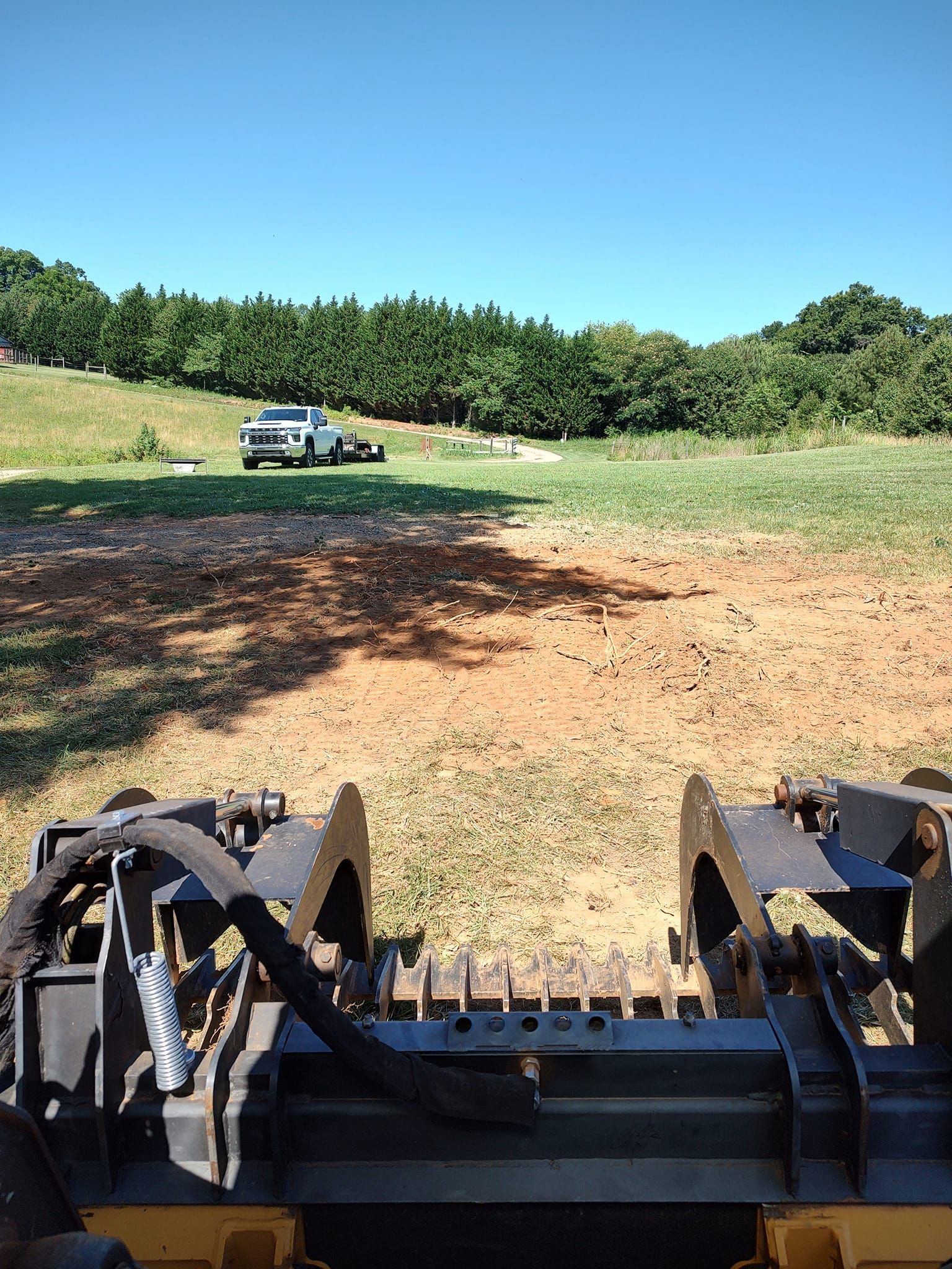 View from a front loader over a dirt field toward a parked car and trees under a clear blue sky