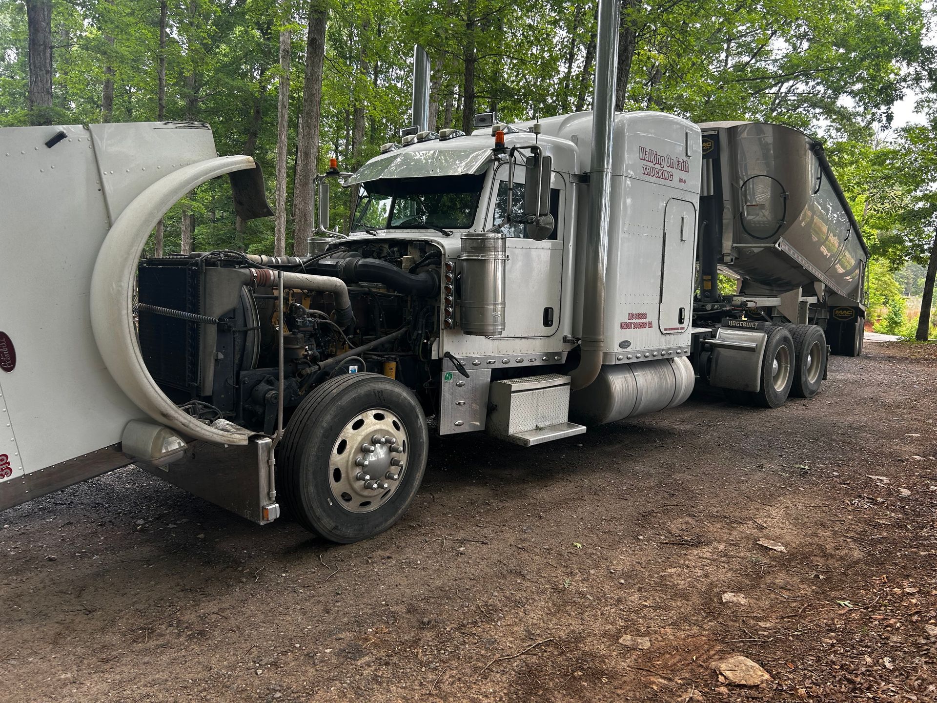 A semi truck with its hood open is parked in a dirt lot.