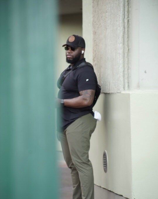 Man in black shirt and cap, khaki pants, and earbuds stands near a wall.