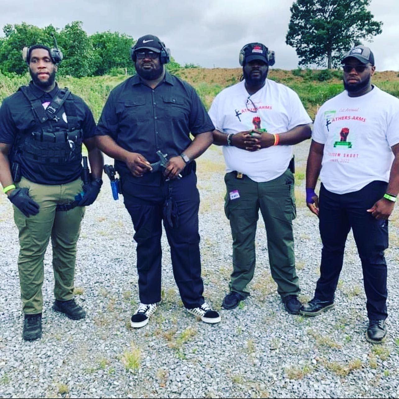 Four people standing outdoors wearing ear protection; two holding firearms. They are wearing various dark colored clothes, near a grassy field.