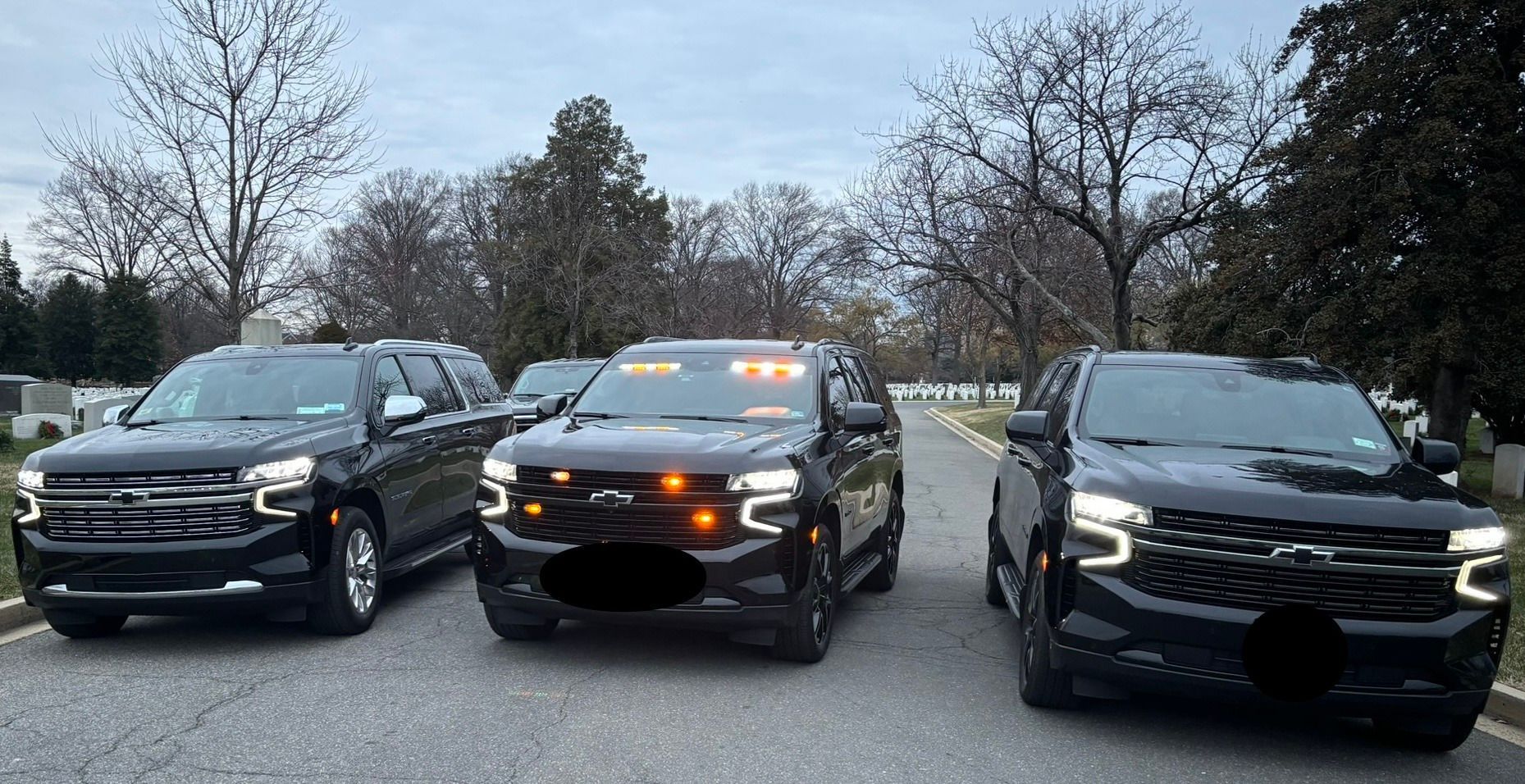 Three black Chevrolet Suburbans parked on a road, some with emergency lights.