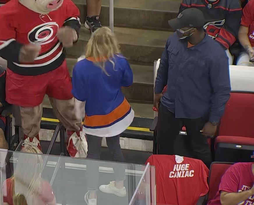 A fan in an Islanders jersey near the ice with a mascot and a man in a black hat.