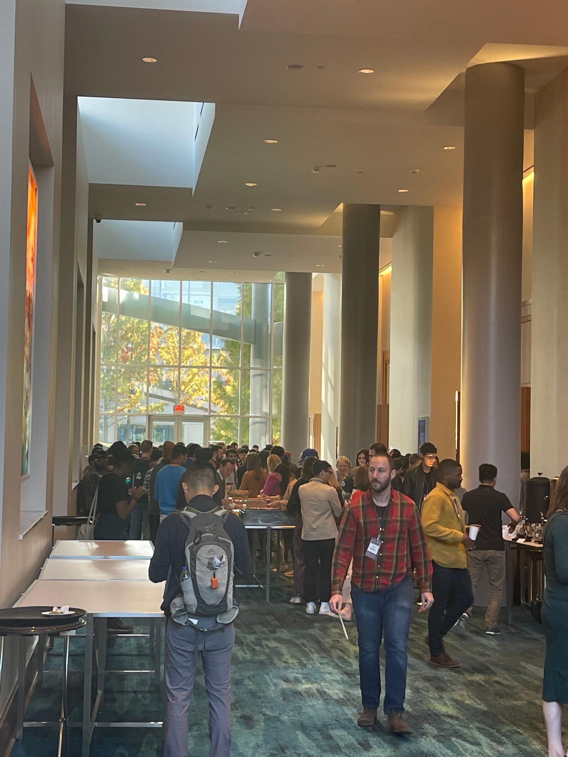 Large hall with people, tables, and columns. Sunlight streams through large windows.