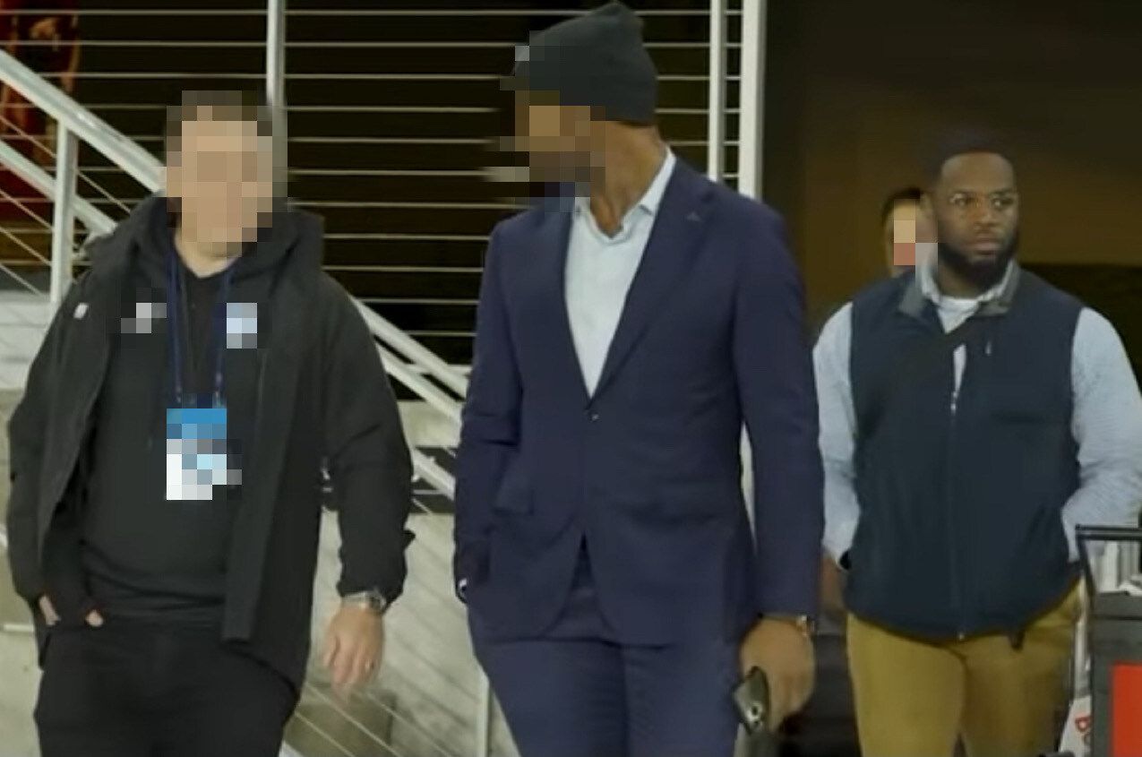 Three men walk in a building. One wears a blue suit, the others wear a black beanie, a vest and jacket.