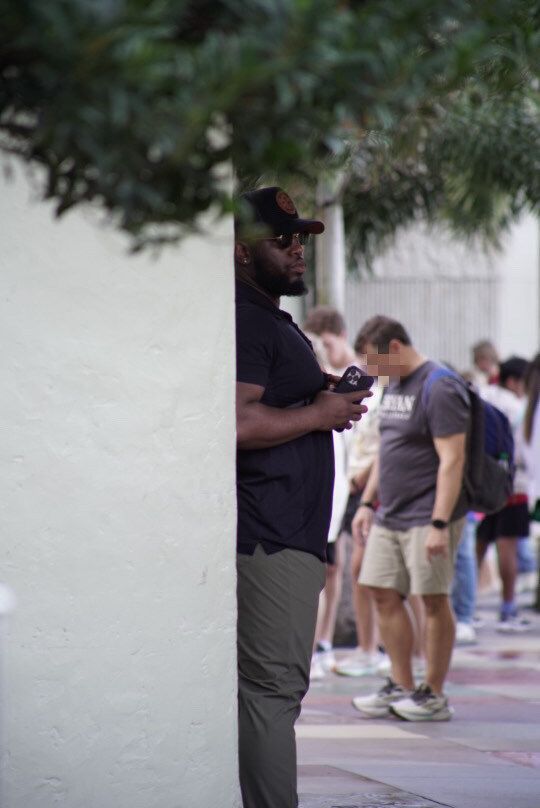 Man in black shirt and cap using a phone, partially obscured by a wall, watching a line of people outdoors.