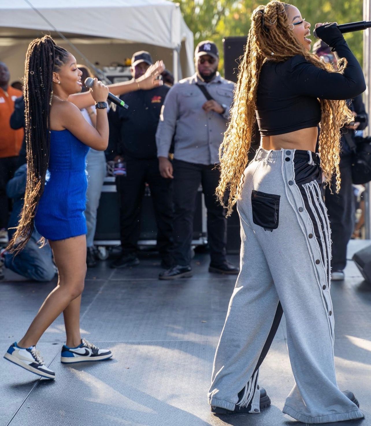 Two women singing on stage. One in blue dress, the other in black top and gray pants. Both holding mics.