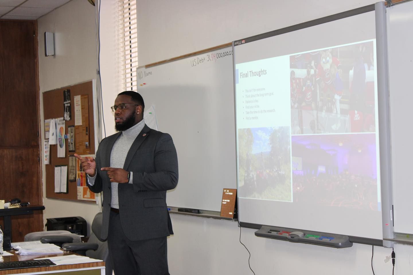 Man in suit presenting to class, pointing at projector screen showing images and text.