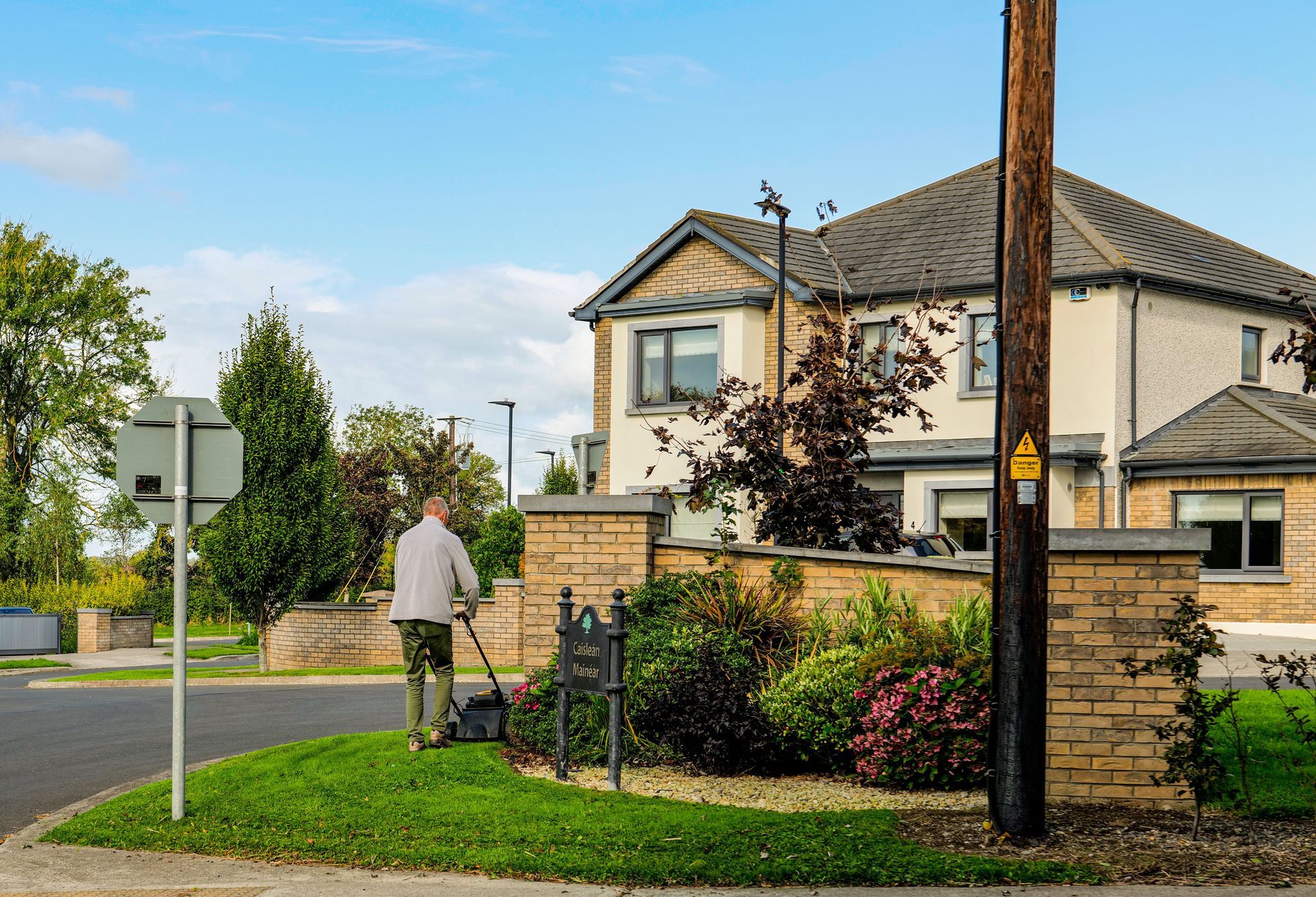 Person walking on a suburban sidewalk beside a two-story house and garden on a sunny day