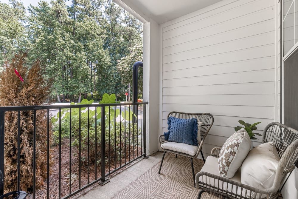 A cozy balcony with two wicker chairs adorned with cushions and a throw pillow, set against a railing overlooking a scenic view of lush green trees. A patterned outdoor rug covers the floor, completing this serene outdoor sitting area at Marq Perimeter in Atlanta, GA.