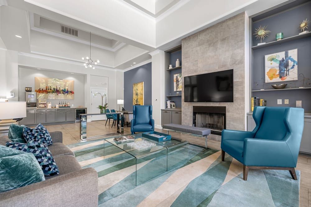 A spacious, modern clubhouse featuring a neutral color palette with blue accents. The room includes a plush beige sofa with patterned blue pillows, two striking blue armchairs, and a glass coffee table on a multi-toned blue area rug. The back of the room has a dining area with a dark blue wall and an elegant table set. A large flat-screen TV hangs above a sleek fireplace, flanked by built-in shelves with decorative items and artworks. High ceilings with recessed lighting and a contemporary chandelier contribute to the room's bright and airy ambiance at Marq Perimeter in Atlanta, GA.