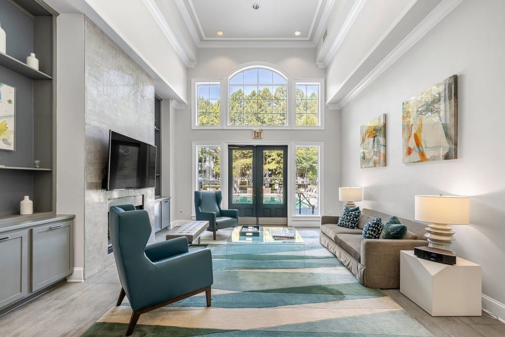 Elegant clubhouse with high ceiling and large arched window allowing natural light to fill the space. The room features a beige L-shaped sofa with decorative pillows, two modern teal armchairs, and a wall-mounted flat-screen TV. A grey and teal area rug anchors the space, and abstract artworks hang on the wall, complementing the room's contemporary aesthetic. The room opens up to an outdoor area through double glass doors at Marq Perimeter in Atlanta, GA.