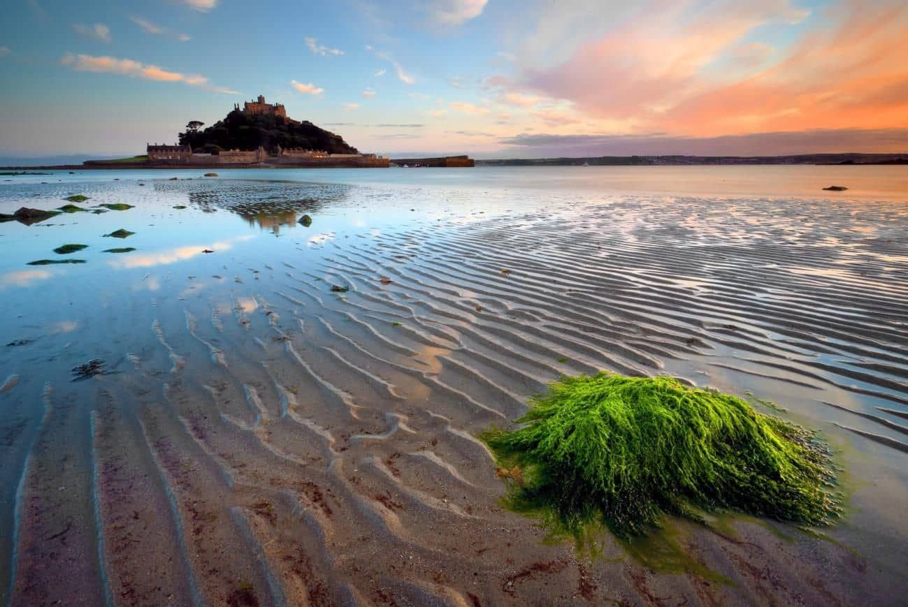 Coastal view of St. Michael's Mount at sunset, with rippled sand, vibrant seaweed, and colorful sky reflections.