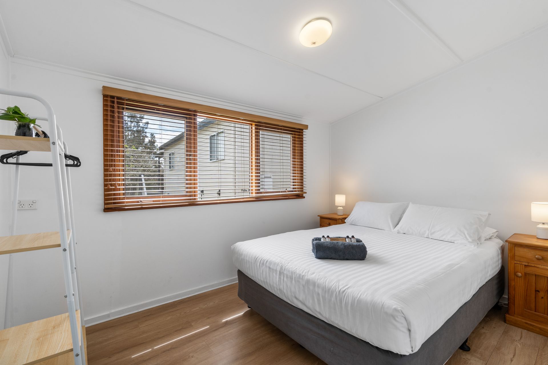A Bedroom With A Queen Bed Featuring Linens, A Wooden Nightstand, And Wooden Blinds — SeaChange Parks Milton in Milton, NSW