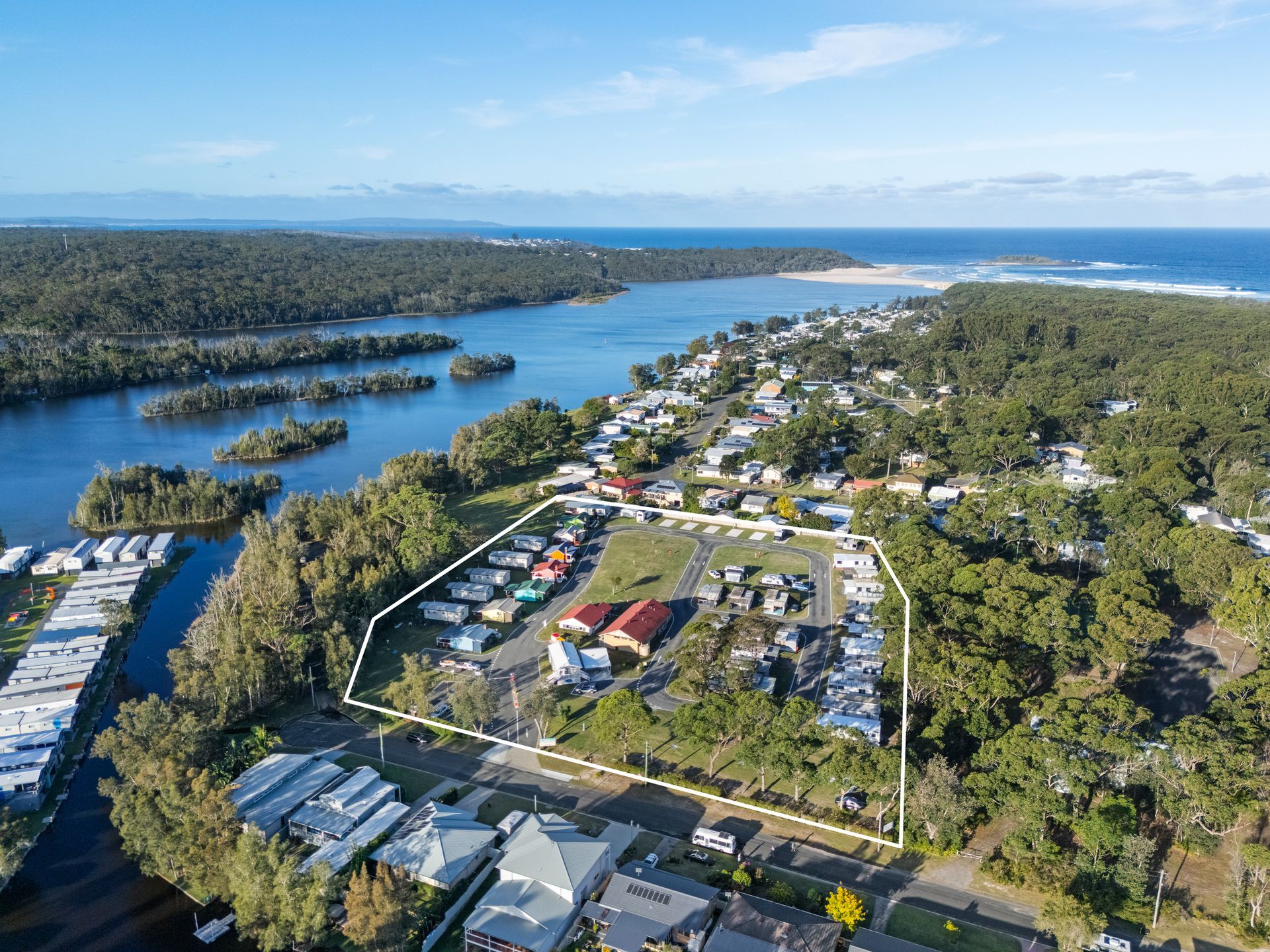 Aerial view: Campground outlined, near water and houses, with green trees, blue sky — SeaChange Parks Milton in Milton, NSW