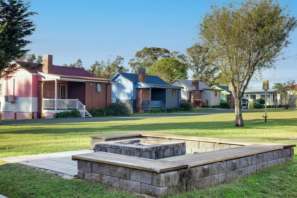 Colourful Cottages Line a Green Lawn with A Stone Fire Pit in The Foreground — SeaChange Parks Milton in Lake Conjola, NSW