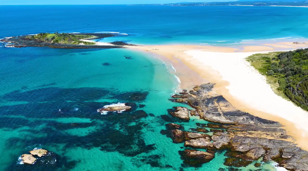 An Aerial View Of A Sandy Spit Connecting A Small, Lush Island To A Sunlit Beach — SeaChange Parks Milton in Milton, NSW