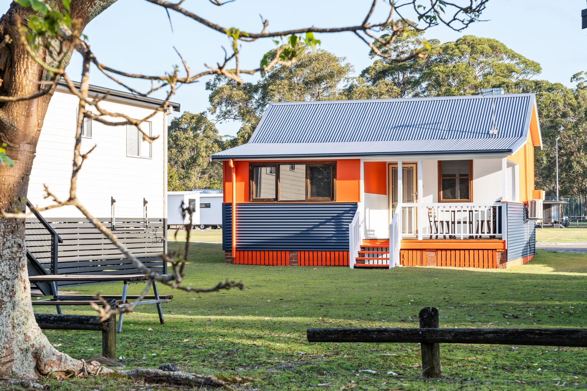 A Small Cabin with Blue and Orange Siding and A Metal Roof Sits on A Grassy Field — SeaChange Parks Milton in Milton, NSW