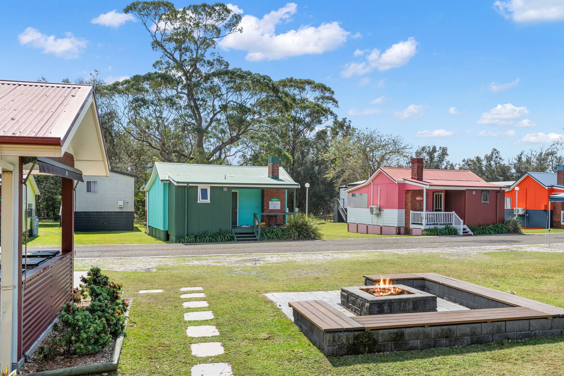 A Sunny Outdoor Area Featuring Small, Colourful Cabins with A Stone Fire Pit — SeaChange Parks Milton in Milton, NSW
