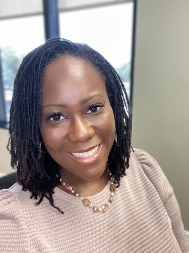 Woman with dark locs smiles at the camera, wearing a light pink sweater and beaded necklace.