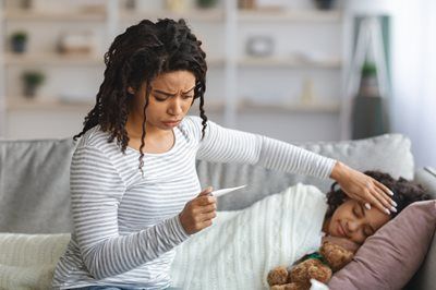 Woman taking child's temperature on a couch; worried expression, child resting with a teddy bear.