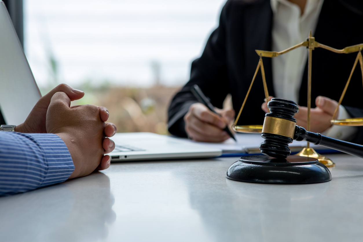 A legal meeting with hands clasped, a gavel, scales of justice, a laptop, and a person writing.