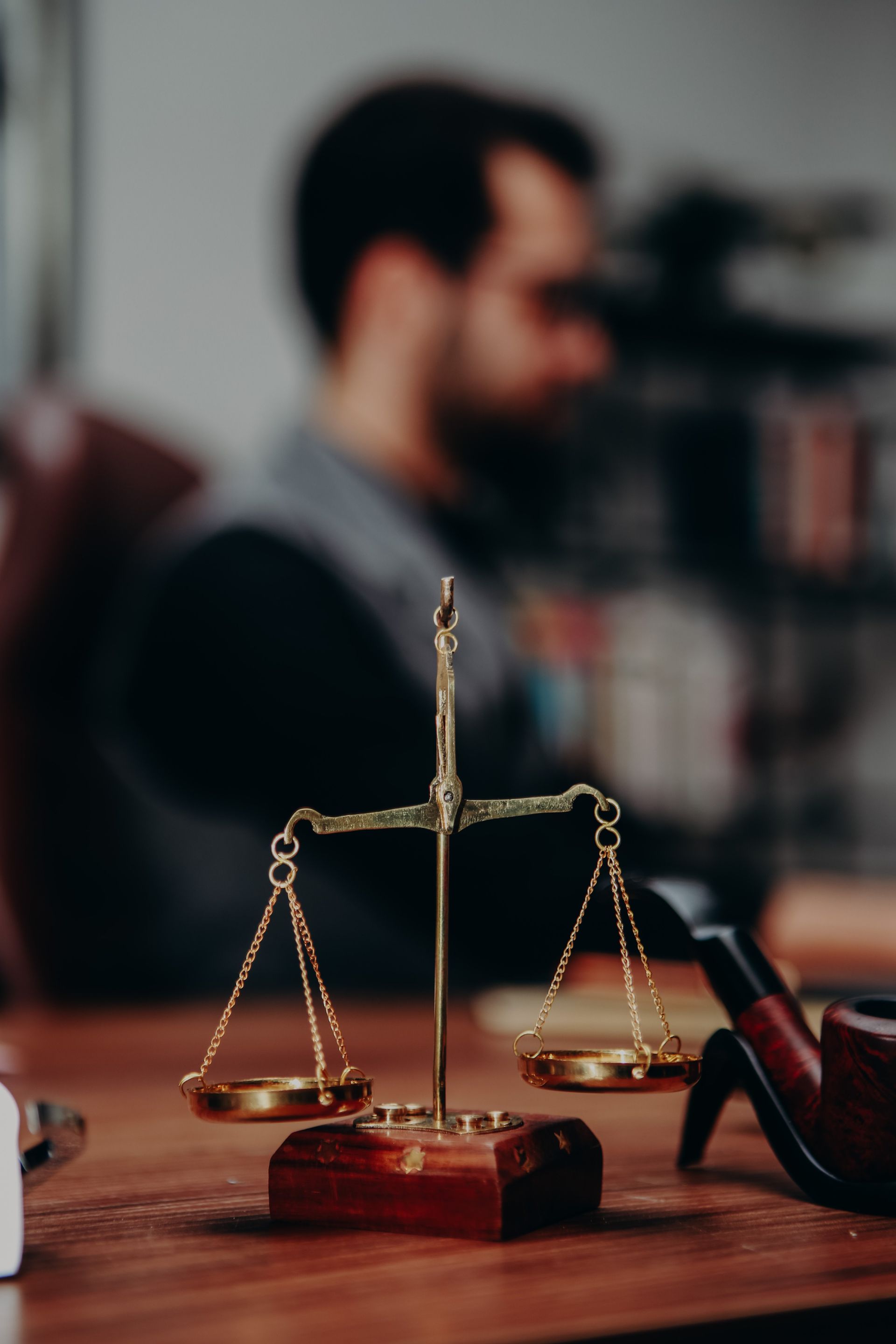 Brass scales of justice on a wooden desk, with a blurred person and office shelves in the background