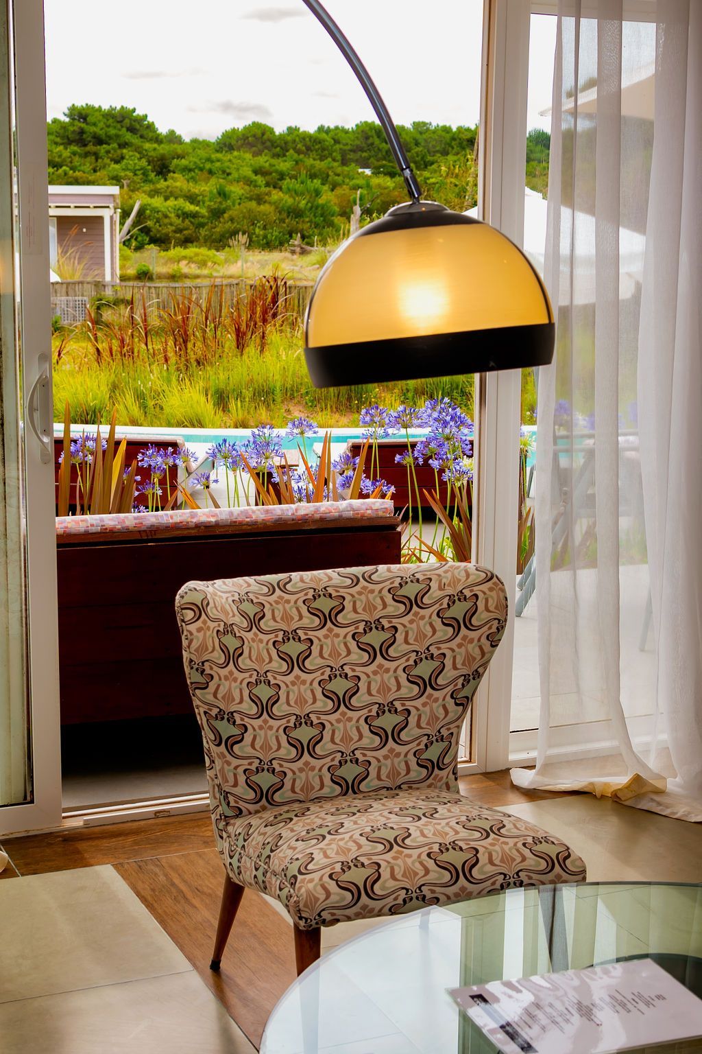 Silla con tela estampada frente a una mesa de cristal, con vista a un patio exterior y vegetación.