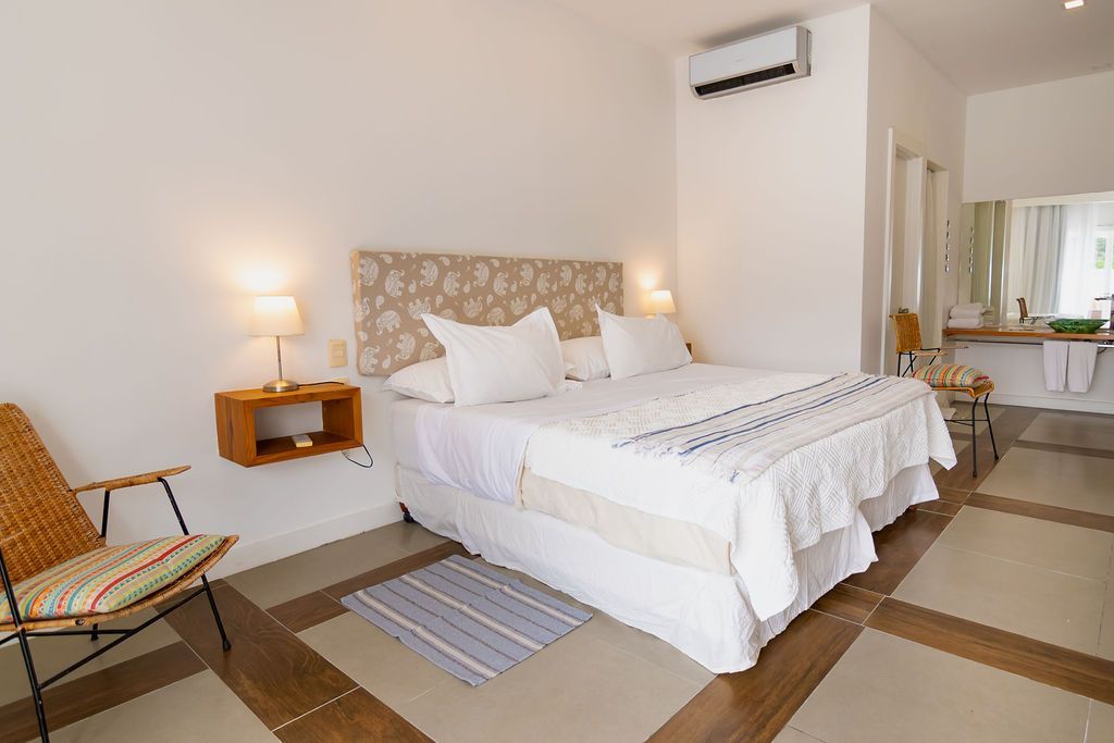 Dormitorio con cama blanca, mesita de noche flotante, silla tejida y puerta abierta a un baño.