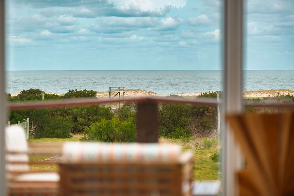Vista del océano desde el interior, a través de una ventana. Se ven el cielo nublado, la playa y la barandilla de un balcón.