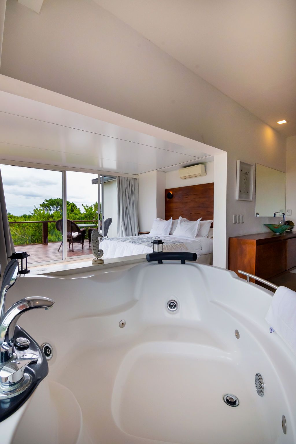 Jacuzzi blanco en primer plano, que conduce a un dormitorio con grandes ventanales, cama y terraza con mesa.