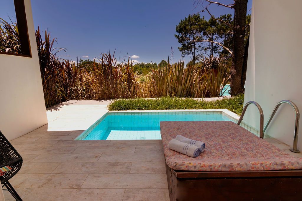 Patio con pequeña piscina, tumbona y césped alto. Cielo azul y árboles al fondo.