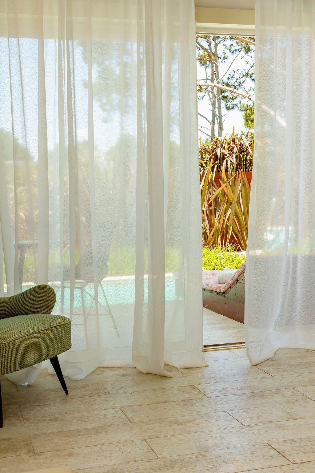 Cortinas blancas transparentes enmarcan una vista de una piscina y follaje, con una silla estampada a la izquierda.