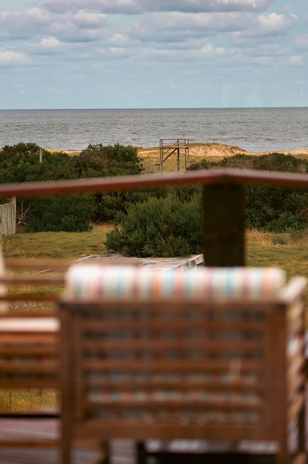 Vista del mar desde una terraza, con sillas de madera en primer plano y arbustos y arena en el medio.