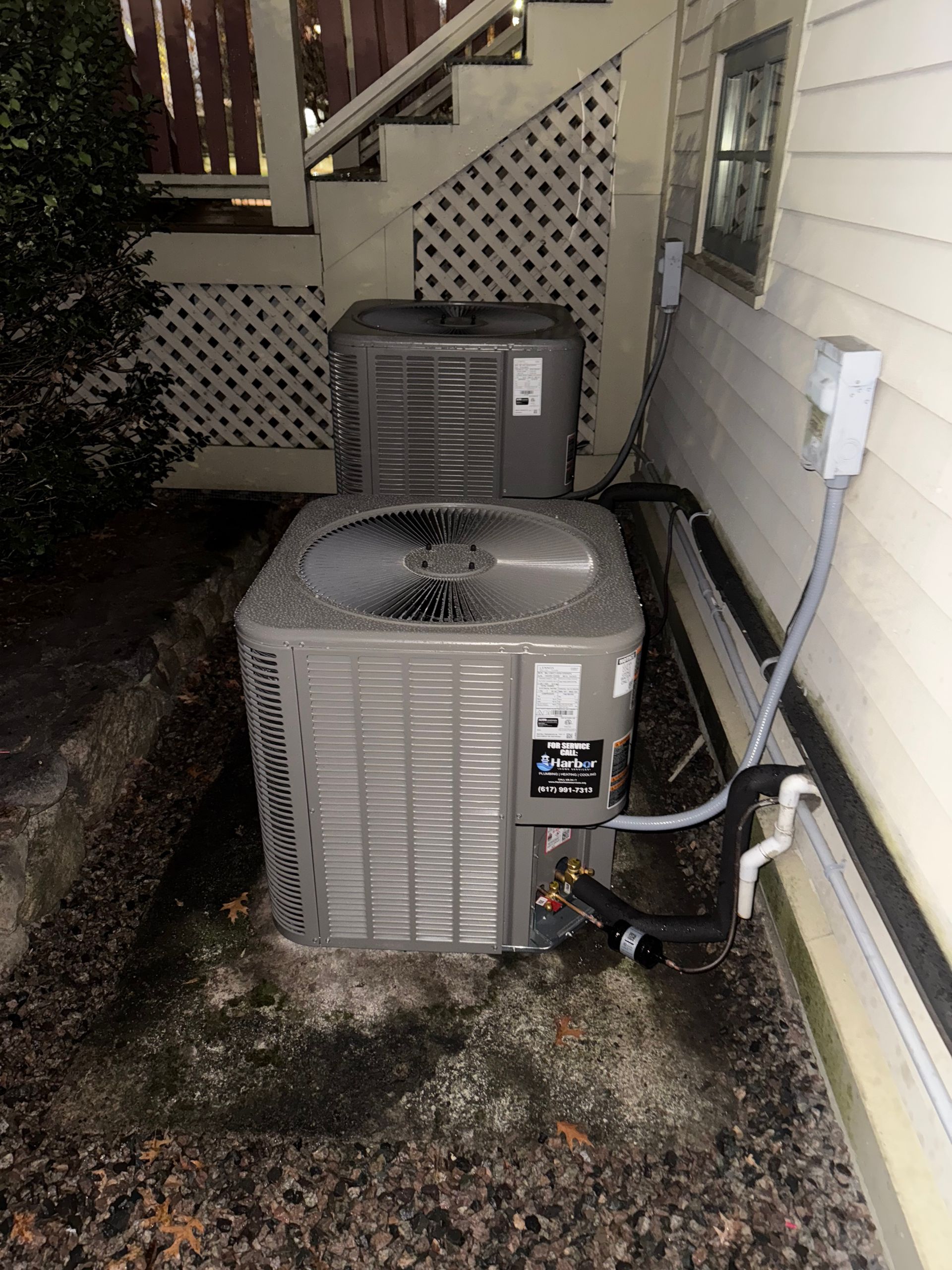 Two outdoor HVAC units beside a house with a lattice porch and visible cables.