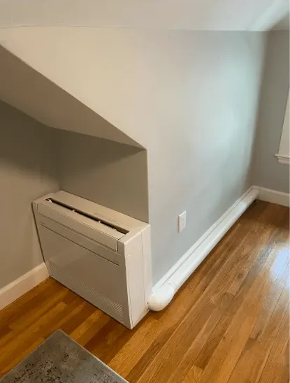 White HVAC unit installed in a corner, below a sloped ceiling, next to wooden flooring.