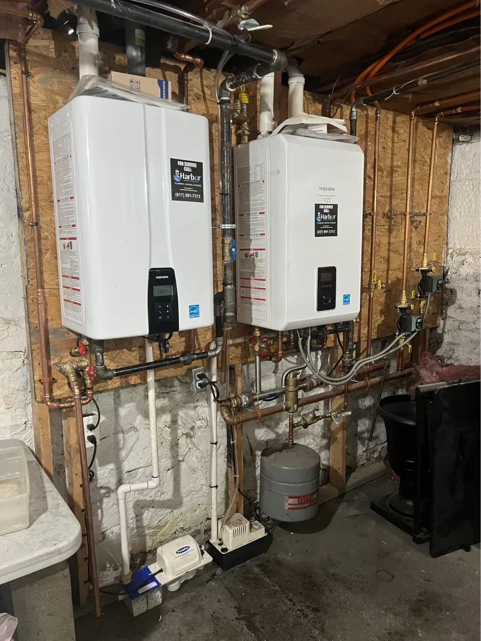 Two white water heaters mounted on wooden paneling with copper pipes in a basement.
